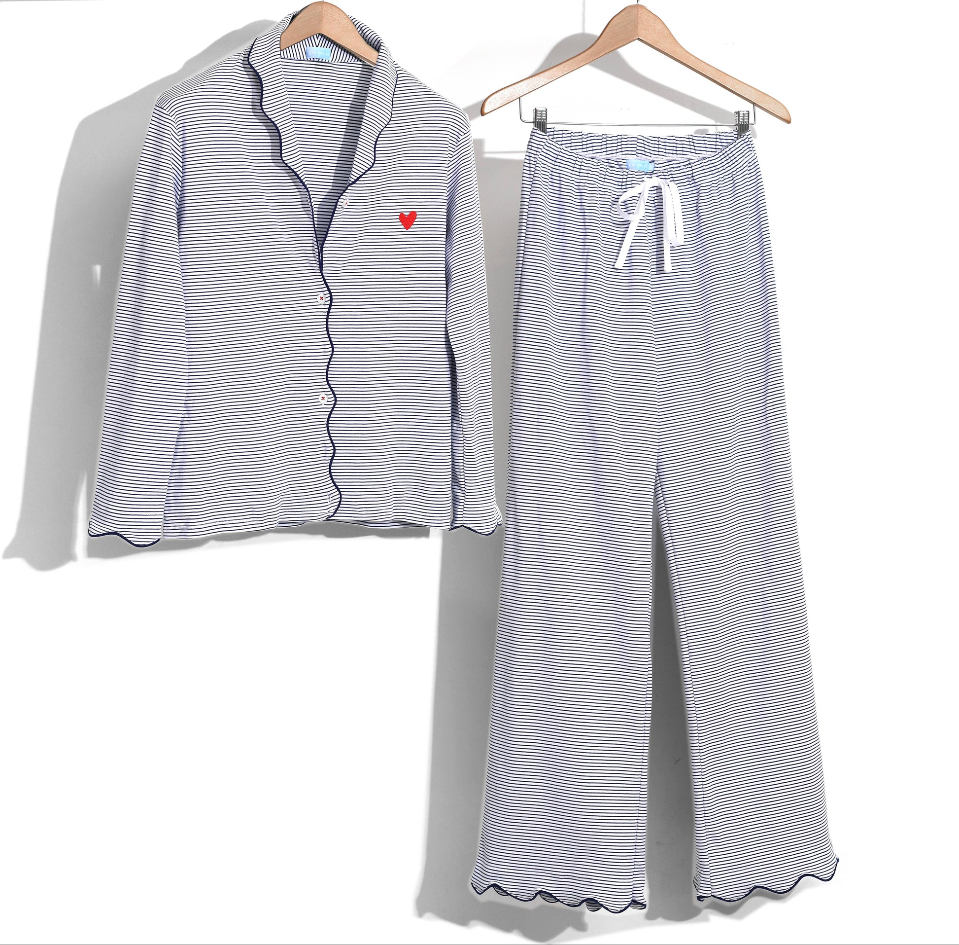 A two-piece women's pajama set with a long-sleeve button-front top and matching wide-leg pants, featuring fine blue and white stripes, scalloped edges, and a small red heart on the chest. The set is displayed on wooden hangers against a white background.